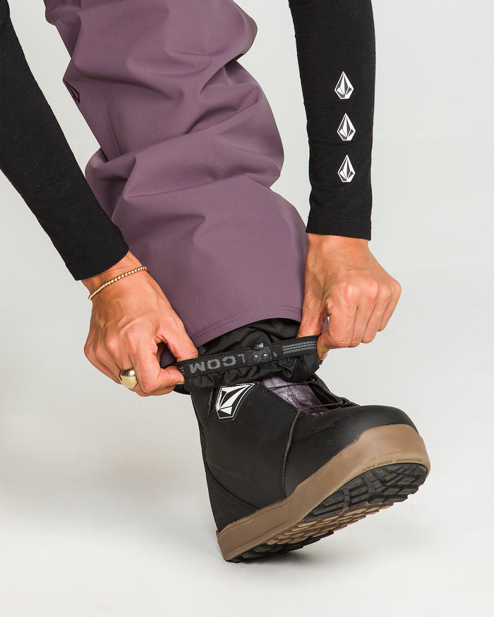 A person wearing Volcom Women’s Stoned Pants in purple adjusts the cuff over a black Volcom snowboarding boot with a brown sole. The individual’s hands hold the pant cuff, and the Volcom logo is visible on both the boot and the sleeve.