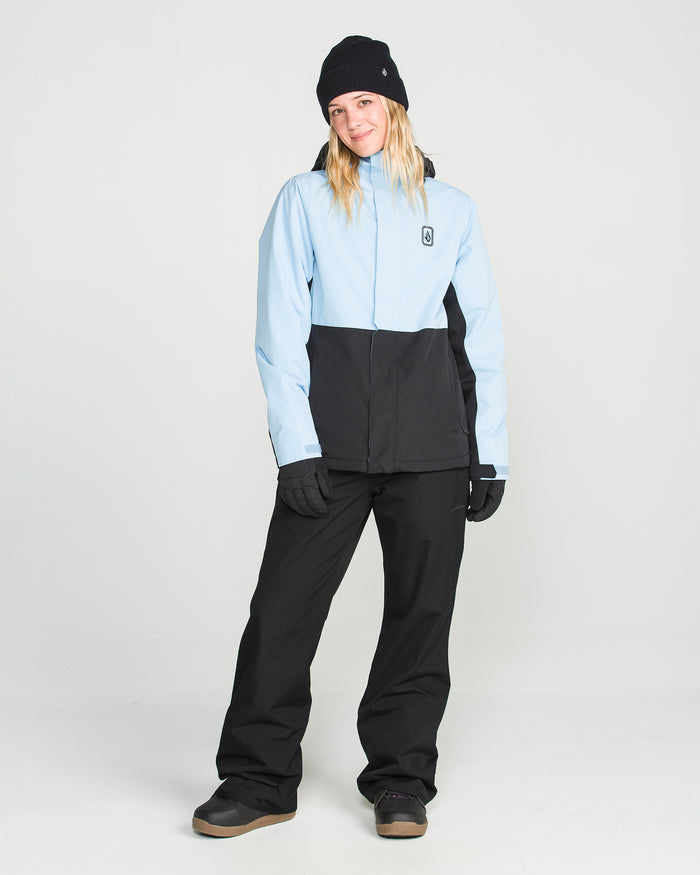 A person wearing black Volcom Women’s Stoned Pants, a light blue and black winter jacket, gloves, boots, and a black beanie stands in a studio with a white background, looking at the camera and smiling slightly.