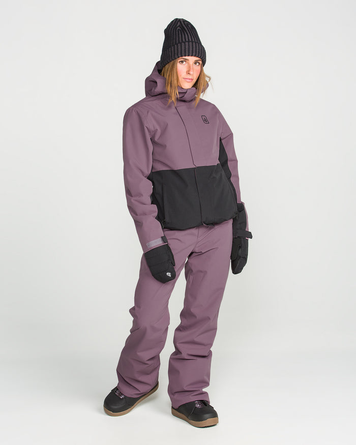 A person stands wearing a purple and black winter snowsuit, Volcom Women’s Stoned Pants, black gloves, black boots, and a knitted black beanie, posing against a plain white background.