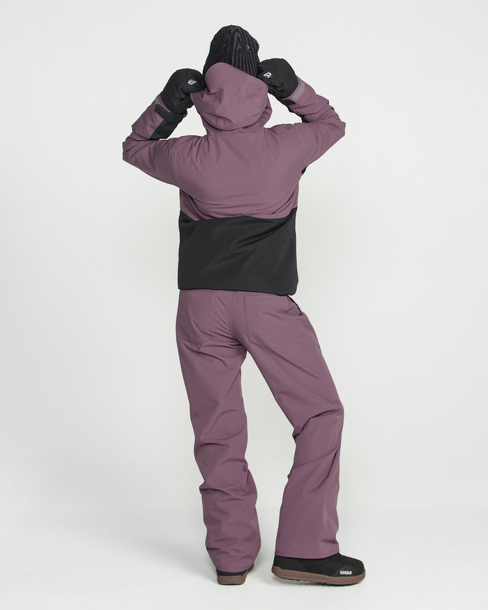 A person stands facing away against a plain white background, wearing a purple and black hooded jacket, black gloves, black shoes, and Volcom Women’s Stoned Pants.