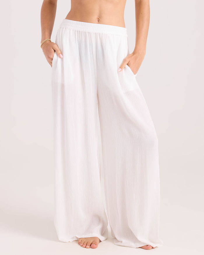 A person stands barefoot, hands in pockets, wearing Volcom Women's Stoneshine Junki Pant—loose, high-waisted, wide-leg white pants in lightweight fabric. Image is cropped at the waist and ankles.