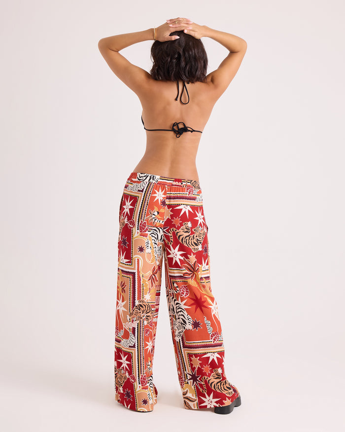 A person stands facing away with arms raised and hands clasped on their head, wearing a black bikini top and Volcom Women's Stoneshine Junki Pants—loose, colorful wide-leg pants featuring red, orange, and white designs with an elastic waistband—paired with black platform shoes against a plain background.