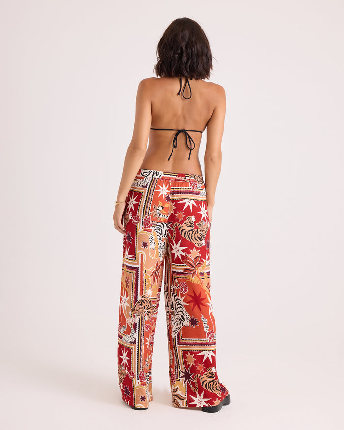 A woman with shoulder-length dark hair stands facing away, wearing a black halter bikini top and the Volcom Women's Stoneshine Junki Pants. The high-waisted, wide-leg pants feature a red, white, and orange floral and geometric pattern and offer a relaxed fit with an elastic waistband for added comfort.