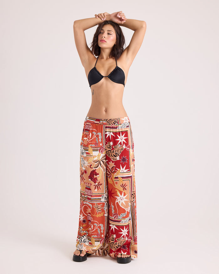 A woman stands with arms raised, wearing a black bikini top and Volcom Women's Stoneshine Junki Pants—relaxed fit, wide-leg pants featuring a red, orange, and white floral patchwork pattern with an elastic waistband—against a plain light background.