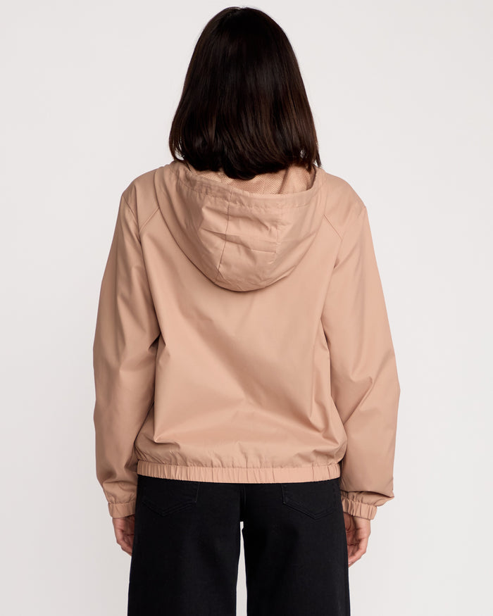 A woman with shoulder-length dark hair is seen from the back wearing a Volcom Women's Stonewind Jacket in beige, featuring a hood and zip front, paired with black pants, standing against a plain white background.