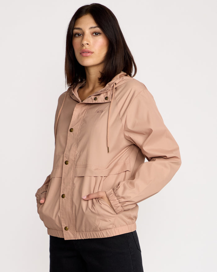 A woman stands with her hands in her pockets, wearing a light pink Volcom Women's Stonewind Jacket featuring a hooded zip front, snap buttons, and front pockets, facing slightly to the side against a plain white background.
