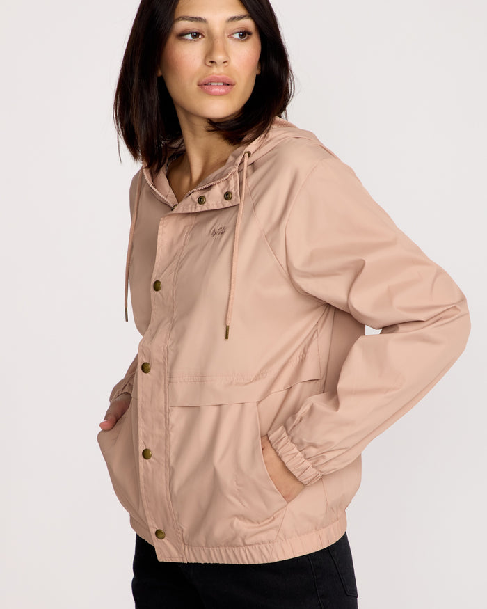 A woman with shoulder-length dark hair wears the Volcom Women's Stonewind Jacket in light pink, featuring a coated taffeta shell and snap buttons, with her hands in the pockets, looking left against a plain light background.