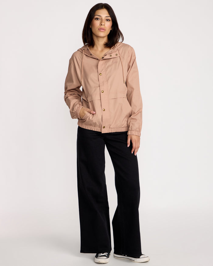 A woman with shoulder-length brown hair and a neutral expression wears the Volcom Women's Stonewind Jacket in light pink, featuring a hooded zip front and coated taffeta shell, paired with black wide-leg pants and sneakers against a plain background.