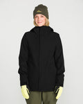 A person wearing the Volcom Women’s Stoney Shadow Insulated Jacket in black, paired with an olive green beanie, olive pants, and light yellow gloves, stands smiling against a plain white background.