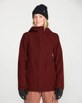 A woman wearing the Volcom Women’s Stoney Shadow Insulated Jacket in maroon, paired with a black beanie and black gloves, smiles at the camera against a plain white background.