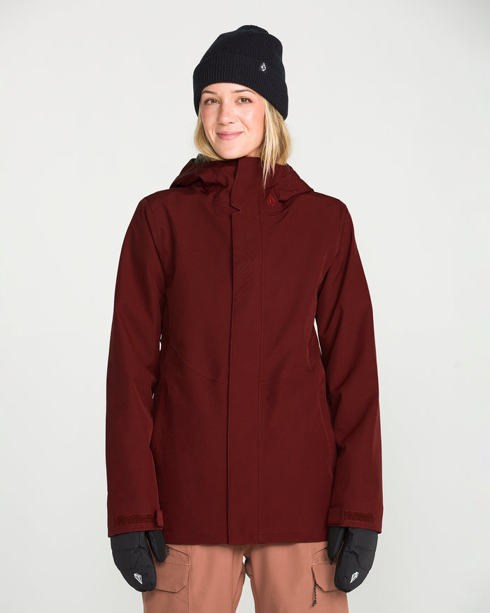A woman wearing the Volcom Women’s Stoney Shadow Insulated Jacket in maroon, paired with a black beanie and black gloves, smiles at the camera against a plain white background.