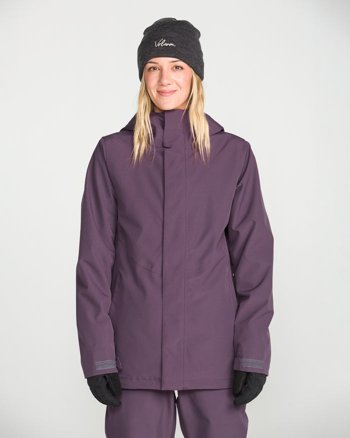 A woman wearing a dark beanie, gloves, and the Volcom Women’s Stoney Shadow Insulated Jacket stands facing the camera with a slight smile against a plain light background.
