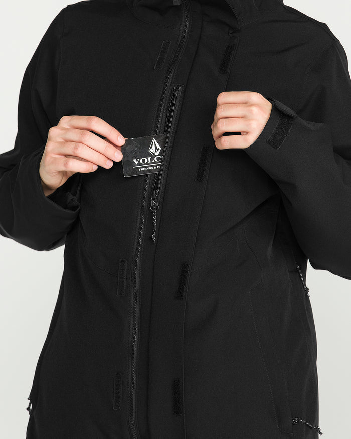 A person is wearing the Volcom Women’s Stoney Shadow Insulated Jacket in black, unzipping it slightly to reveal a patch with the Volcom diamond-shaped logo above the brand name. Only the torso and hands are visible.