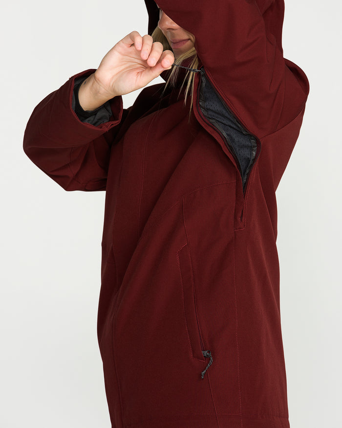 Wearing the Volcom Women’s Stoney Shadow Insulated Jacket in maroon, a person pulls the hood drawstring, partly hiding their face, while a zippered side pocket is visible.