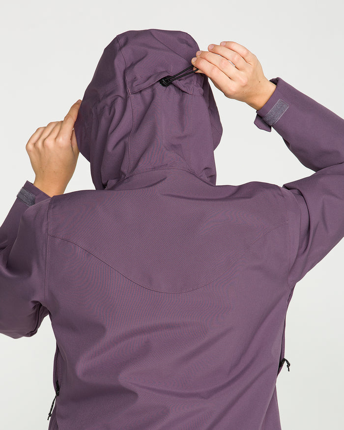 A person wearing the Volcom Women’s Stoney Shadow Insulated Jacket in purple adjusts the hood using a back drawstring, with only their back visible.