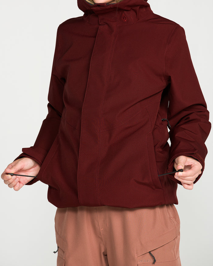 Wearing the Volcom Women’s Stoney Shadow Insulated Jacket in maroon and light brown pants, a person adjusts the jacket’s drawstrings against a plain white background; their face is not visible.