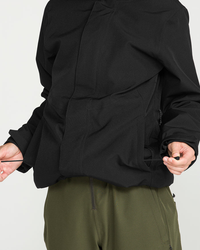 A person wearing the Volcom Women’s Stoney Shadow Insulated Jacket in black, featuring 15K waterproof protection, and olive green pants adjusts the drawstring at the bottom of the jacket. Their head is not visible.