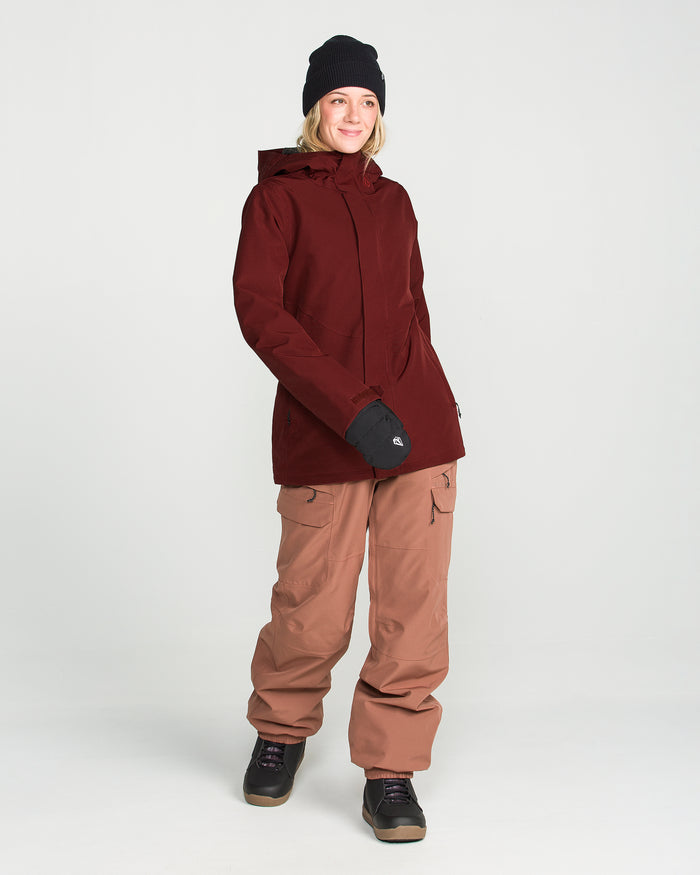 A person wearing the Volcom Women’s Stoney Shadow Insulated Jacket, brown snow pants, black gloves, and a black beanie stands facing forward and smiling against a plain white background.