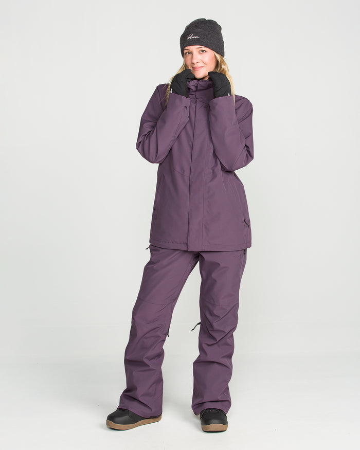 Wearing the Volcom Women’s Stoney Shadow Insulated Jacket along with matching pants, black gloves, black boots, and a gray beanie, a person smiles and poses in front of a plain white background.