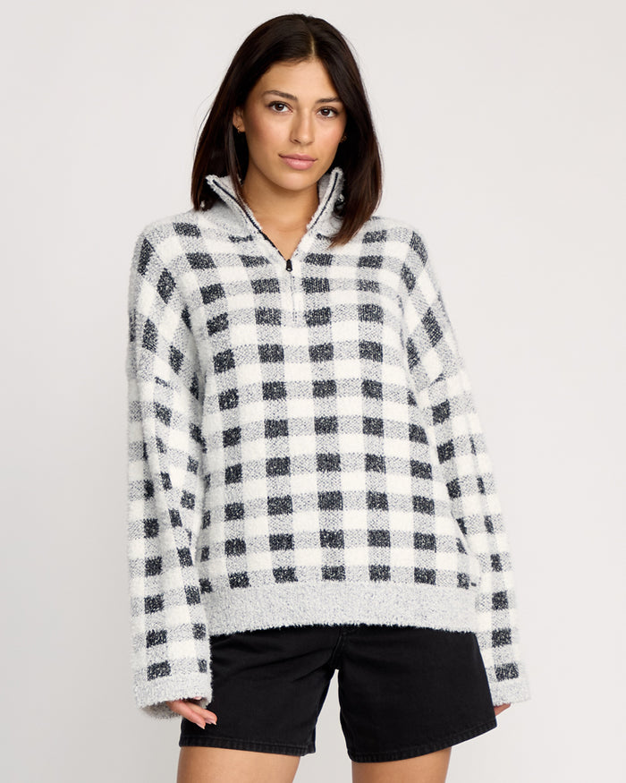 A person with straight, dark hair wears the Volcom Women's Sugarstone Shag Sweater—a white and black plaid pullover with a half-zip collar—paired with black shorts, standing against a plain light background.