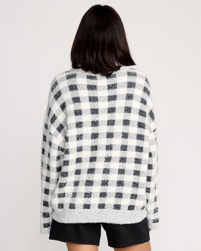 A woman with shoulder-length dark hair faces away from the camera, wearing the Volcom Women's Sugarstone Shag Sweater in black and white plaid with black shorts against a plain light background.