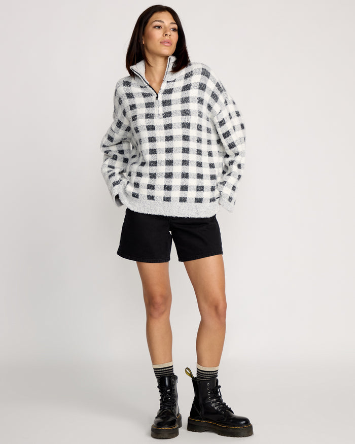 A woman stands against a plain background wearing the Volcom Women's Sugarstone Shag Sweater, black shorts, striped socks, and black lace-up boots. Her hands are in her pockets and she looks slightly to the side.