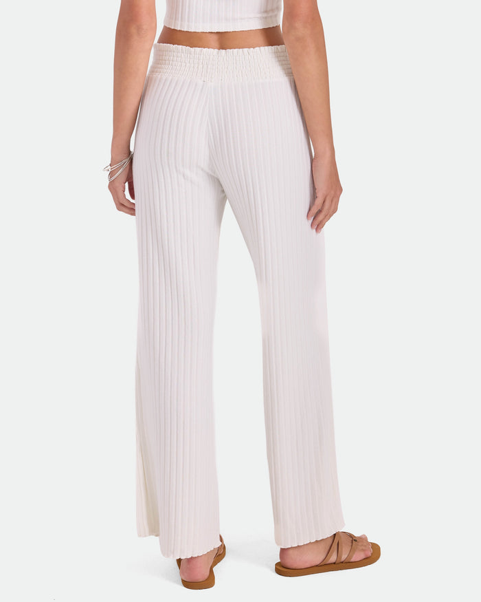 A person is seen from the back wearing Volcom Women's Sun Lounge Pants—high-waisted, wide-leg white pants with a relaxed fit and elastic waistband—paired with brown sandals and a cropped, sleeveless top for an effortlessly comfortable look.