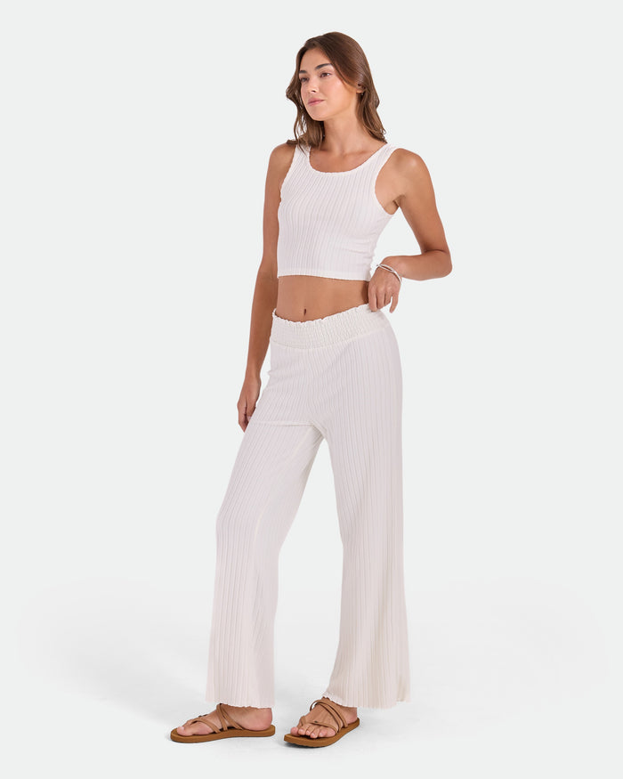 A woman stands in a relaxed pose wearing Volcom Women's Sun Lounge Pants with an elastic waistband and relaxed fit, paired with a sleeveless white crop top and sandals, against a plain light background.