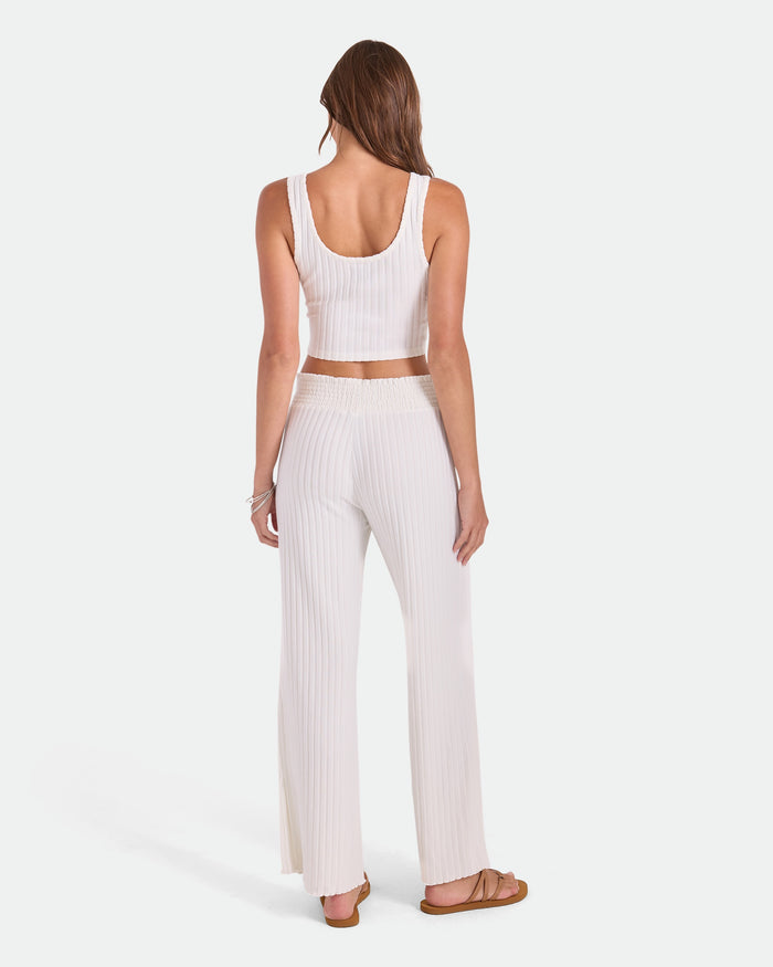 A woman with long brown hair faces away, wearing Volcom Women's Sun Lounge Pants—relaxed fit with elastic waistband—and a white ribbed crop top and sandals, shown on a plain white background.