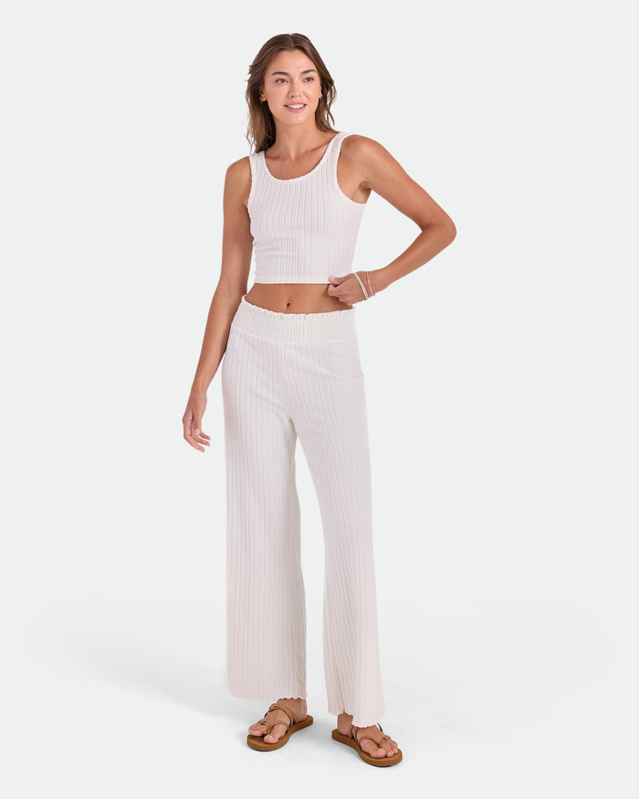 A woman smiles with one hand on her hip, wearing Volcom Women's Sun Lounge Pants—high-waisted, relaxed fit with an elastic waistband—paired with a sleeveless white ribbed crop top, sandals, and bracelets against a plain white background.