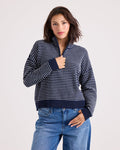 A woman with shoulder-length dark hair wears the Volcom Women's Sun Of Sand Sweater—a navy and white striped quarter-zip design—paired with blue jeans, standing against a plain light background with one hand touching her collar.