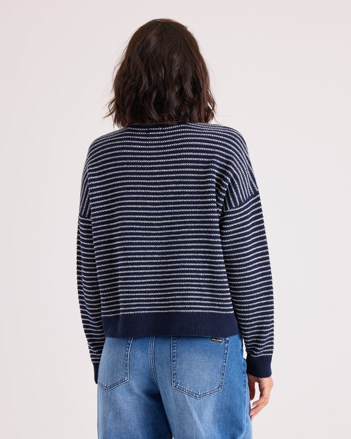 A person with wavy dark hair is shown from the back against a plain light background, wearing the Volcom Women's Sun Of Sand Sweater—a relaxed fit navy blue and white striped pullover—paired with blue jeans.