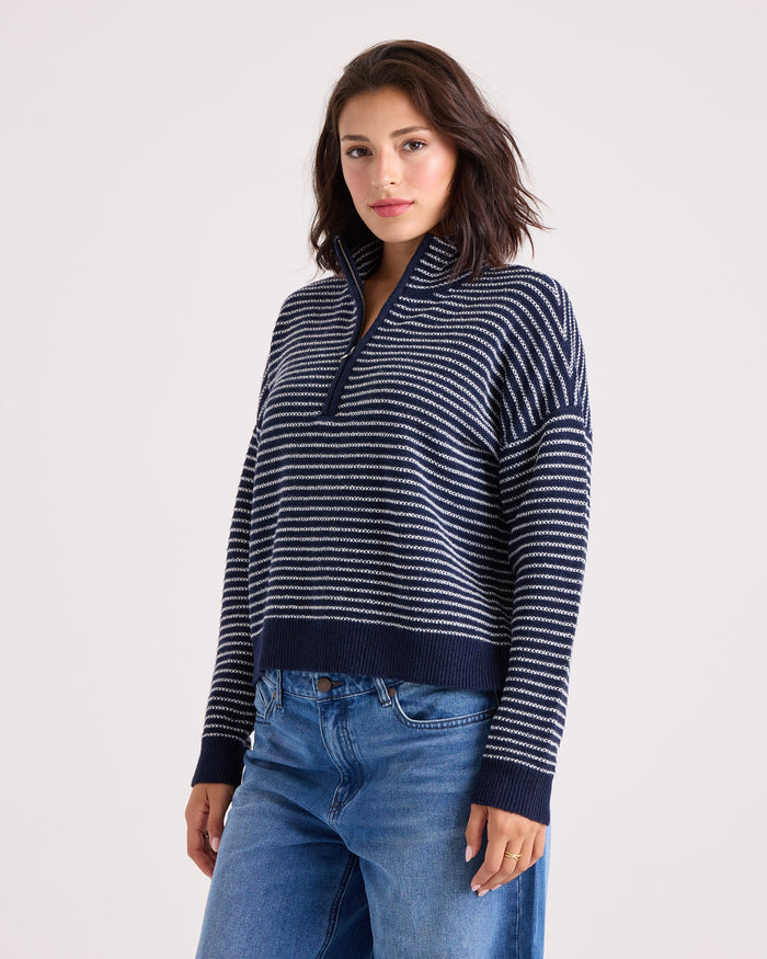 A woman wearing the Volcom Women's Sun Of Sand Sweater—a navy and white striped quarter-zip sweater with a collar—paired with blue jeans, stands against a plain light background, looking at the camera with a neutral expression.