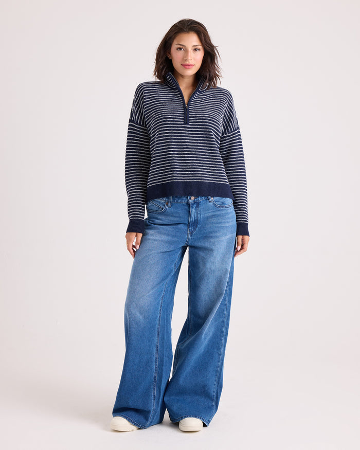 A woman stands against a plain background wearing the Volcom Women's Sun Of Sand Sweater—a navy striped, relaxed fit pullover—paired with wide-leg blue jeans and white sneakers. She looks at the camera with a relaxed expression, her hands by her sides.