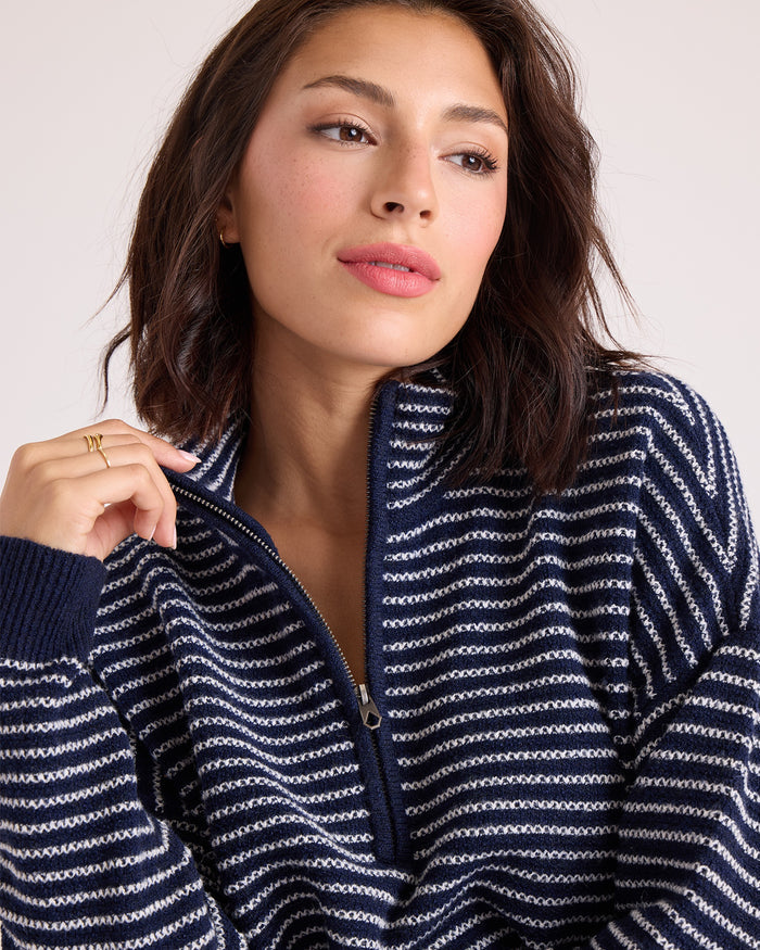 A woman with shoulder-length dark hair wears the Volcom Women's Sun Of Sand Sweater, a navy and white striped quarter-zip with a relaxed fit. The sweater is partially unzipped as she looks slightly to the side with a neutral expression against a plain background.