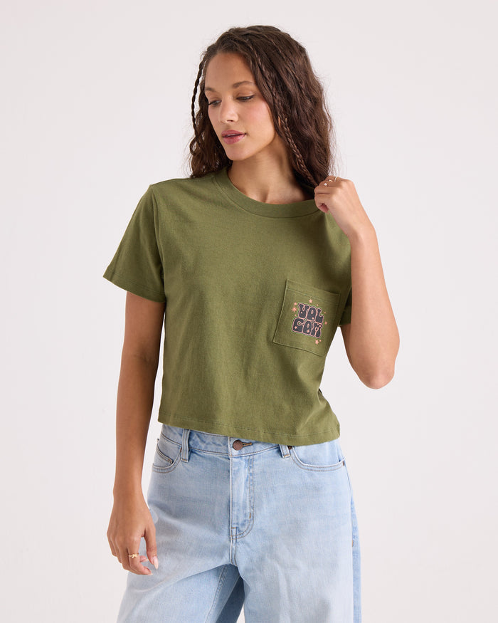 A woman wearing the Volcom Women's Super Melt Pocket Dial Tee in green—made from 100% cotton with a crew neck and pocket—and light blue jeans stands against a plain background, looking down and touching her hair.