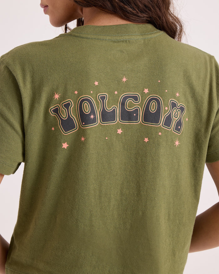 A person with long brown hair is seen from behind, wearing Volcom's Women's Super Melt Pocket Dial Tee—an olive green, 100% cotton crew neck featuring bold, retro-style VOLCOM lettering and small pink stars on the back.