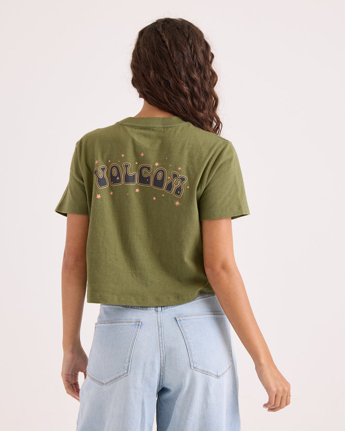 A person with long wavy hair, wearing a green Volcom Women's Super Melt Pocket Dial Tee and light blue jeans, is shown from the back against a plain background.