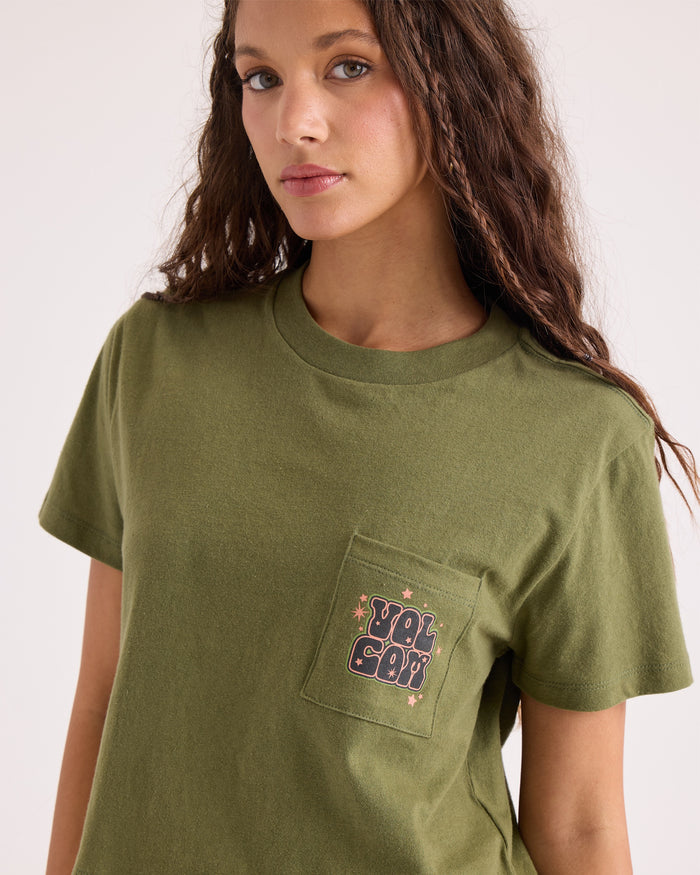 A woman with long curly hair wears the Volcom Women's Super Melt Pocket Dial Tee—an olive green, 100% cotton crew neck featuring a pocket graphic with a patch that reads "YOU GOT" in pink outlined letters surrounded by small stars.