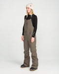 A person stands facing slightly left on a plain white background, wearing a beige beanie, a black long-sleeve top, and the Volcom Women’s Swift Bib in brown, made from recycled fabric.