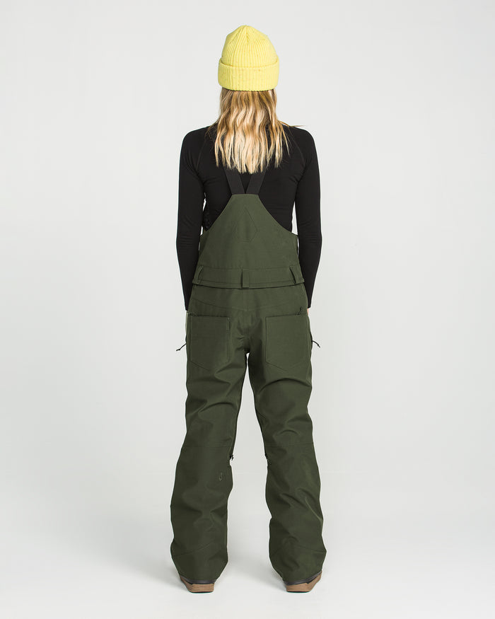 A person stands facing away, wearing a yellow beanie, a black long-sleeve shirt, and the Volcom Women’s Swift Bib—green wide-leg overalls with large back pockets made from recycled fabric. The background is plain white.