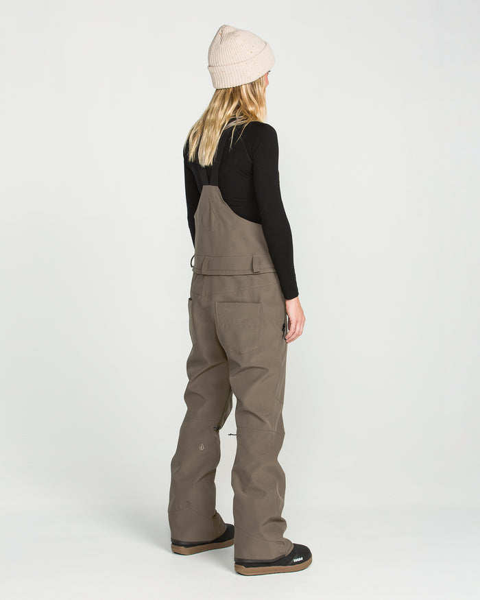 A person with long blonde hair, wearing a beige beanie, black long-sleeve top, and the Volcom Women’s Swift Bib in taupe—crafted from recycled fabric—stands facing away on a plain white background.