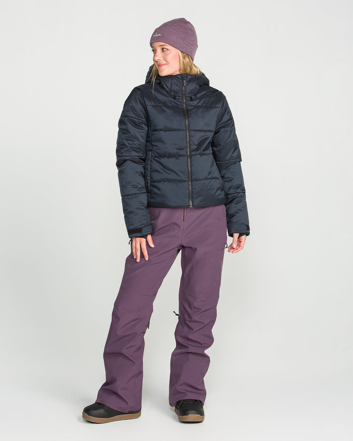 A woman wearing the Volcom Women’s Swift Bib in purple, teamed with a purple beanie, black puffer jacket, and black boots, stands against a plain white background looking to her left with a relaxed posture.