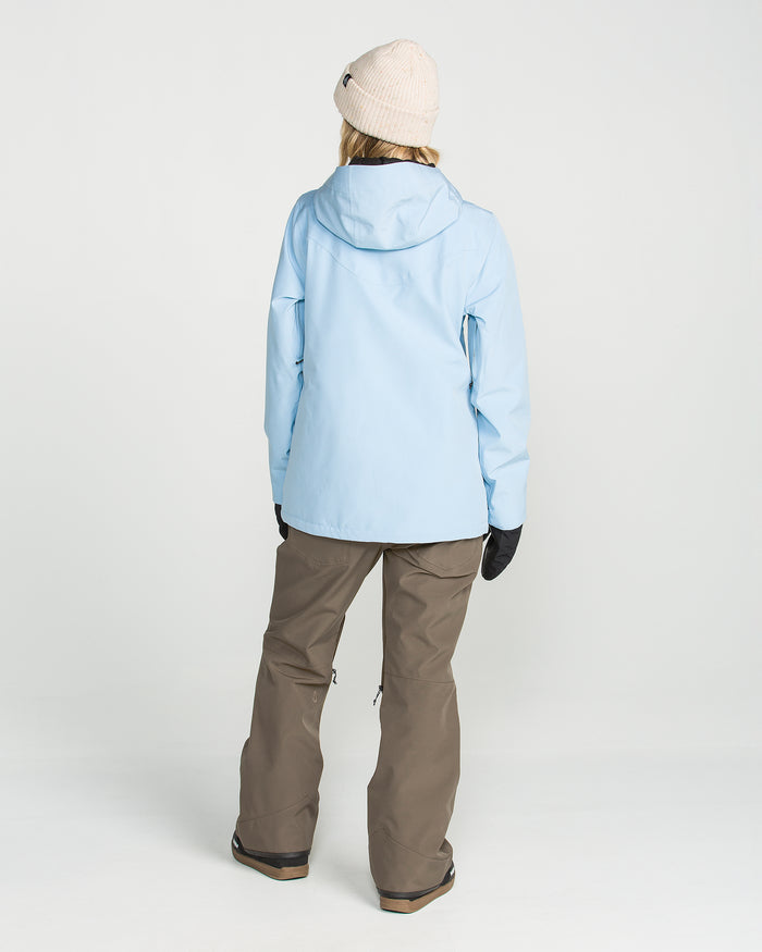 A person is standing facing away against a plain white background, wearing a beige knit hat, light blue hooded jacket, black gloves, and the Volcom Women’s Swift Bib—brown waterproof overalls made from recycled fabric.