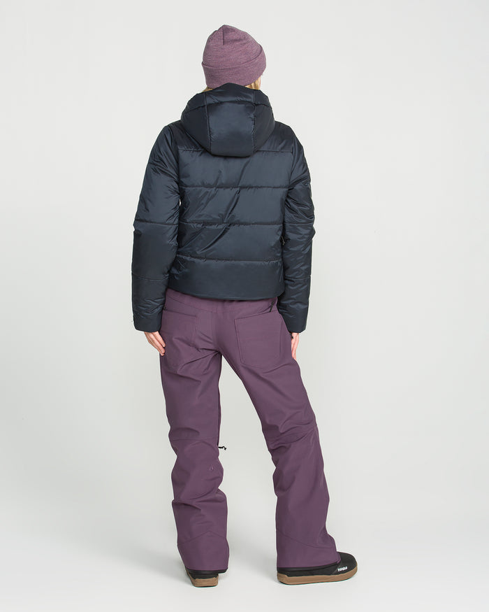 A person stands facing away against a plain white background, wearing a purple beanie, a black puffer jacket, and the Volcom Women’s Swift Bib—purple women’s bib overalls made from recycled fabric.