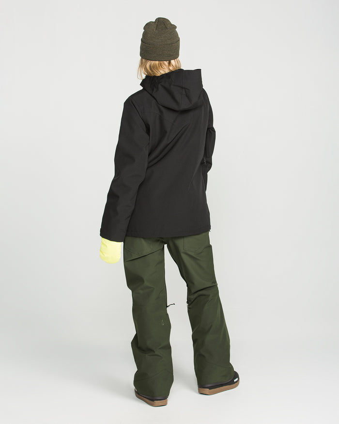 A person stands facing away against a plain white background, wearing the Volcom Women’s Swift Bib in olive green—waterproof snow pants made from recycled fabric—paired with a dark hooded jacket, beanie, and yellow gloves.