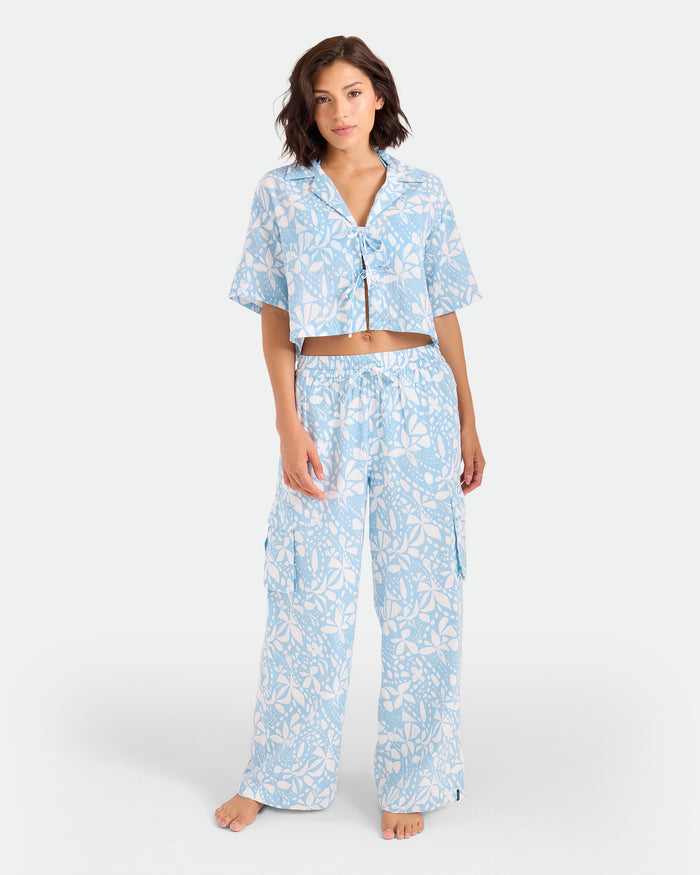 A woman stands barefoot against a white background wearing the Volcom Women's Tied Up Crop Shirt, a light blue and white floral-patterned cotton pajama set with a cropped short-sleeve top and loose wide-leg pants.