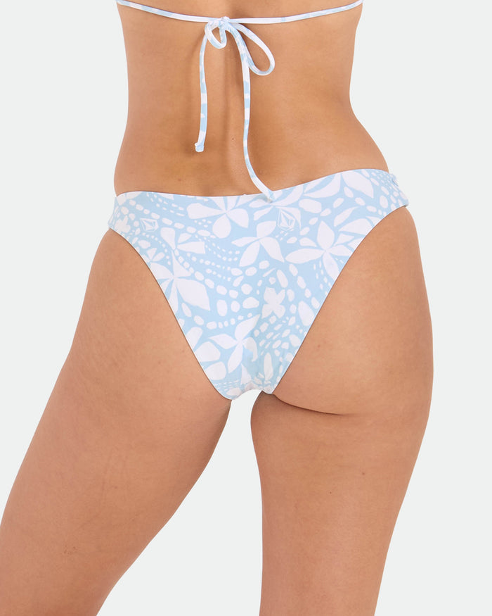 A woman models the Volcom Women's Tribal Stone 90s V Bottom bikini in light blue and white floral print, shown from the back with visible skimpy top ties. The recycled nylon swimwear pops against a plain white background.