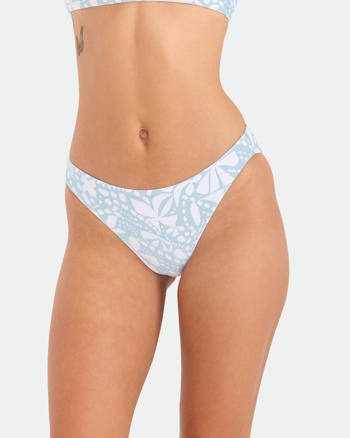 A woman models Volcom's Women's Tribal Stone Hipster Full Bikini Bottom in Summer Song, made from recycled nylon; she stands against a plain white background, showing her midsection, upper thighs, and a small tattoo near her waist.