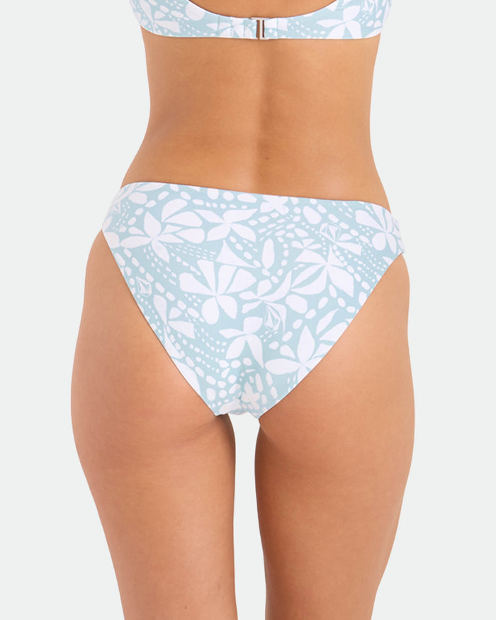 A close-up rear view shows a person wearing Volcom’s Women's Tribal Stone Hipster Full Bikini Bottoms in Summer Song—a light blue, recycled nylon swimwear with white abstract florals—against a plain background.